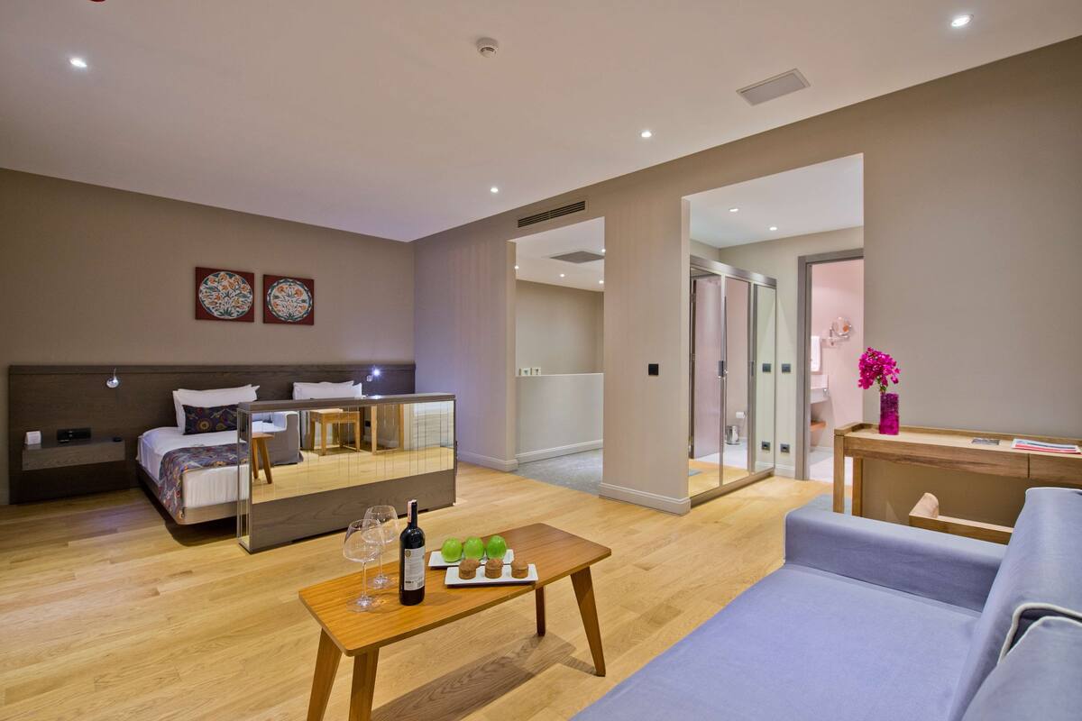 Una habitación de hotel moderna con una cama, una mesa de madera con una botella y vasos, un sofá y un espejo que refleja la habitación.