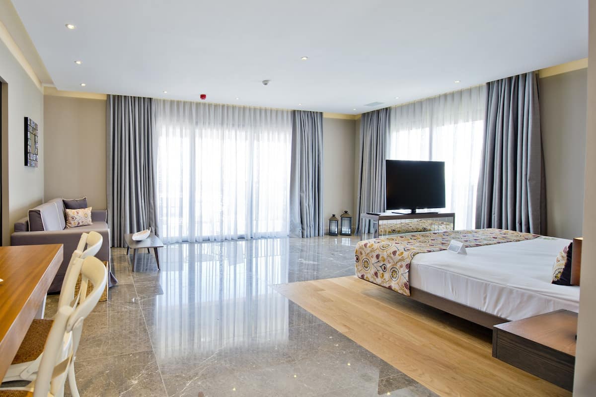 Una habitación de hotel moderna con una cama grande, un televisor de pantalla plana, una zona de comedor con mesa y sillas, y ventanas amplias con cortinas.