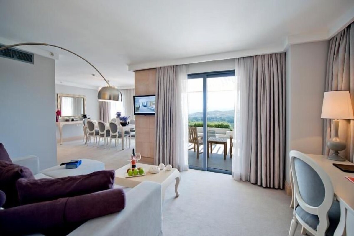 Habitación de hotel con sofá, comedor con sillas, televisor y vista a través de las cortinas.