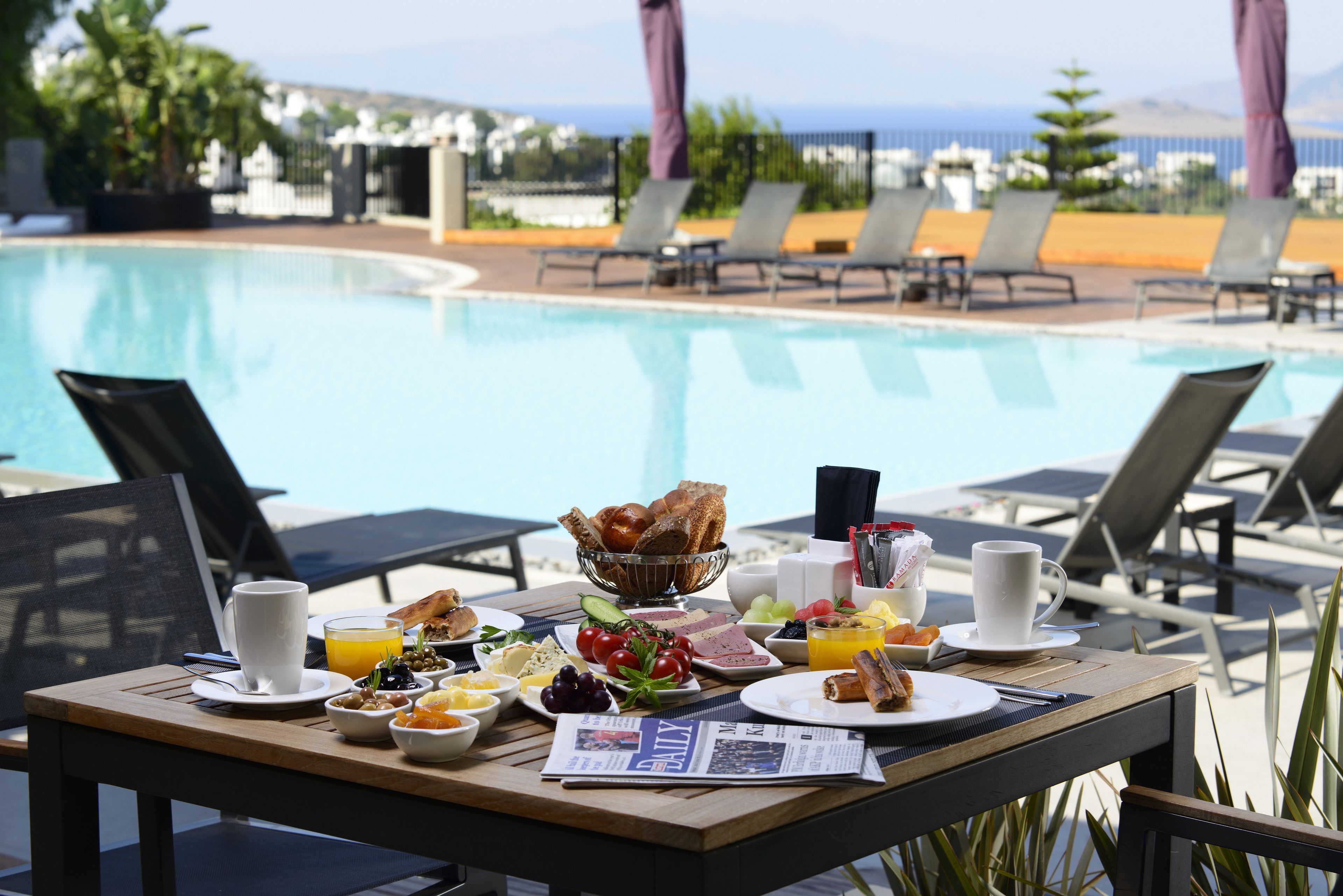 Una mesa de desayuno con diversos alimentos y un periódico, con vistas a una piscina con sillas de descanso y sombrillas.