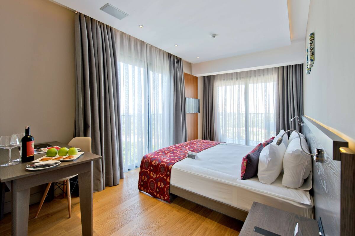 Habitación de hotel con cama, escritorio, silla, mesa con copas de vino y manzanas, y cortinas.