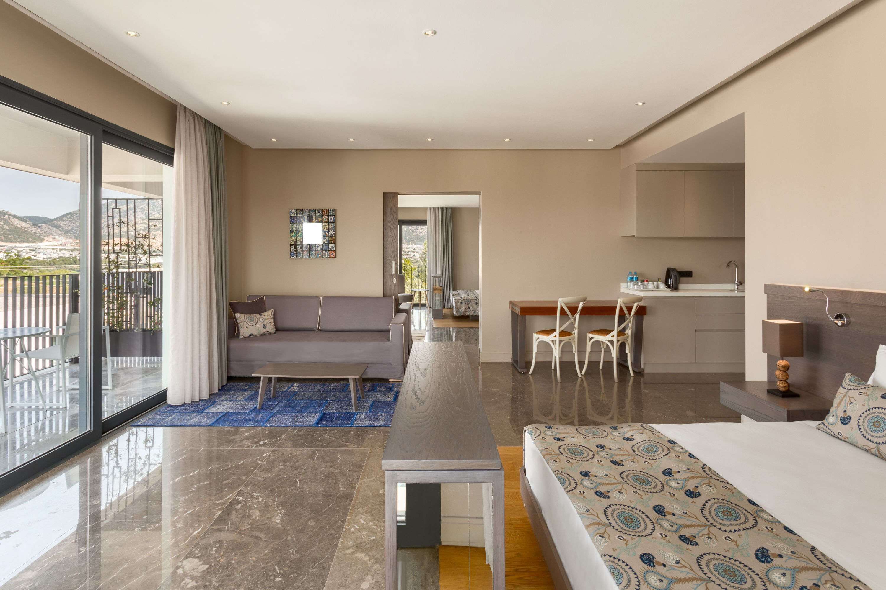 A modern hotel room with a large bed, a desk, a sofa, and a balcony with a view of the mountains.