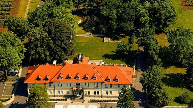 Vista aérea de una extensa propiedad con un edificio destacado de techo rojo, rodeada de exuberante vegetación y amplios jardines.