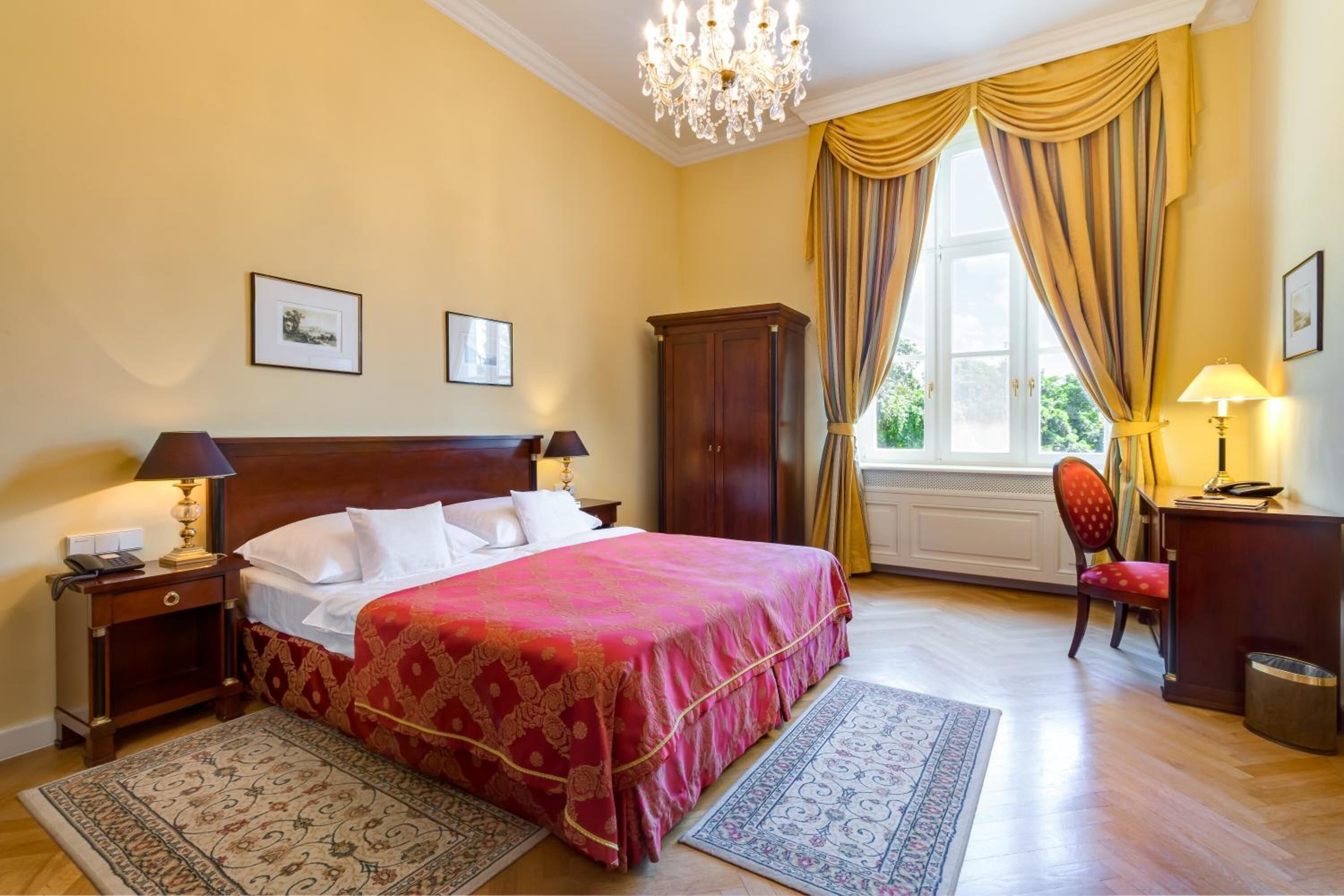 A bedroom with a large bed, a wooden wardrobe, a desk with a lamp, and a window with curtains.