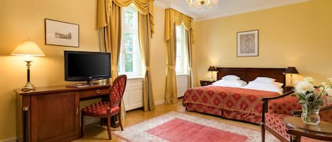 Una habitación de hotel con escritorio de madera, televisor, cama con cubrecama rojo estampado y candelabro.