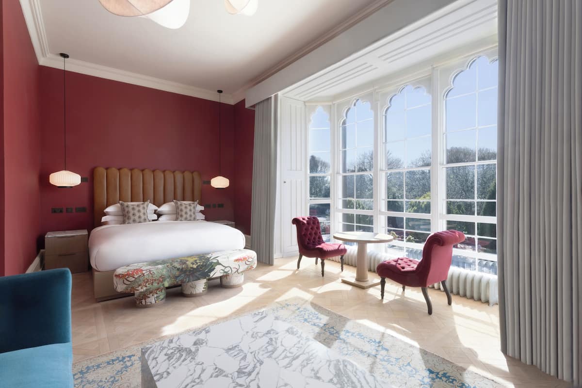 A bedroom with a large bed, a small table, two chairs, and a view of the outdoors through large windows.