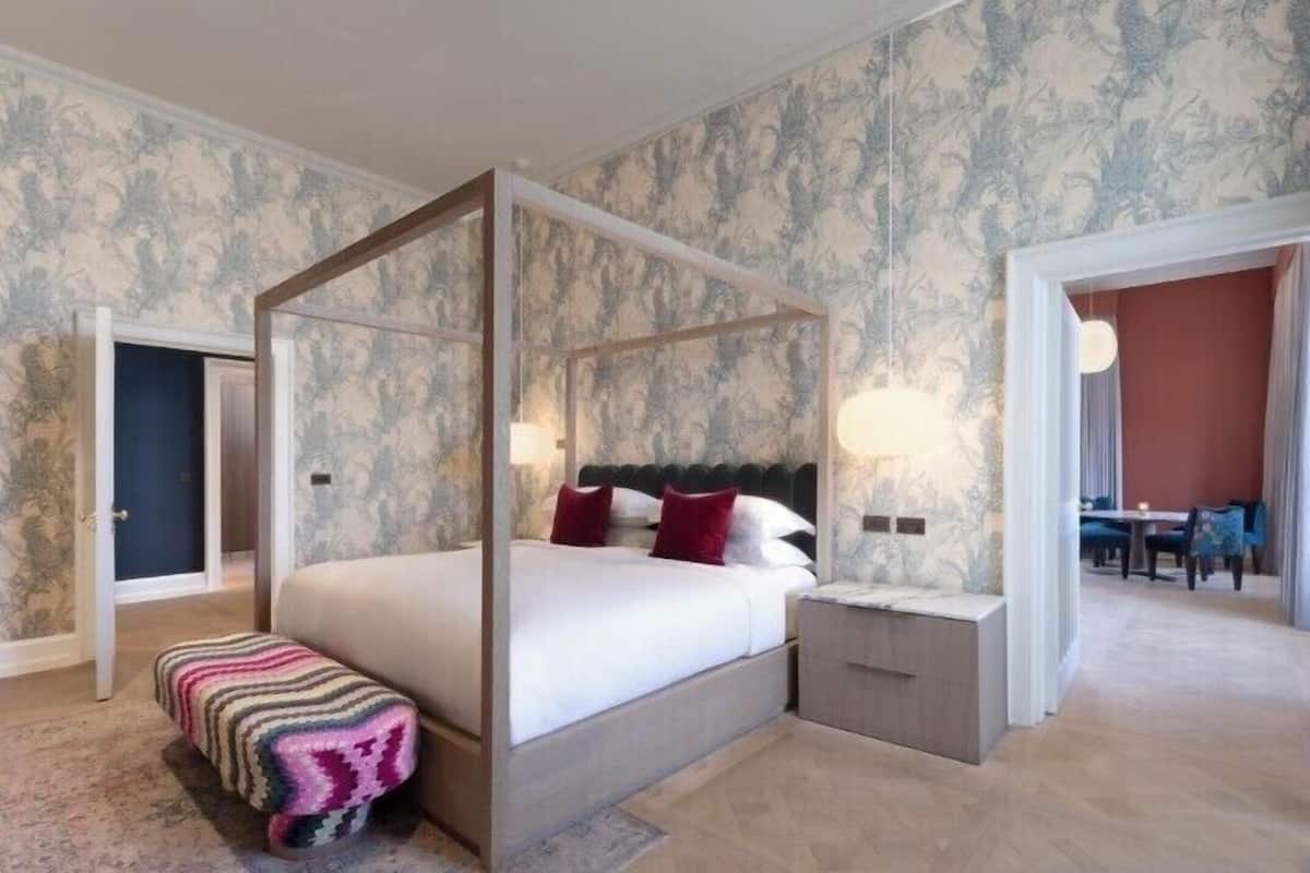 A four-poster bed with a nightstand, a bench, and a dining area visible in the background.