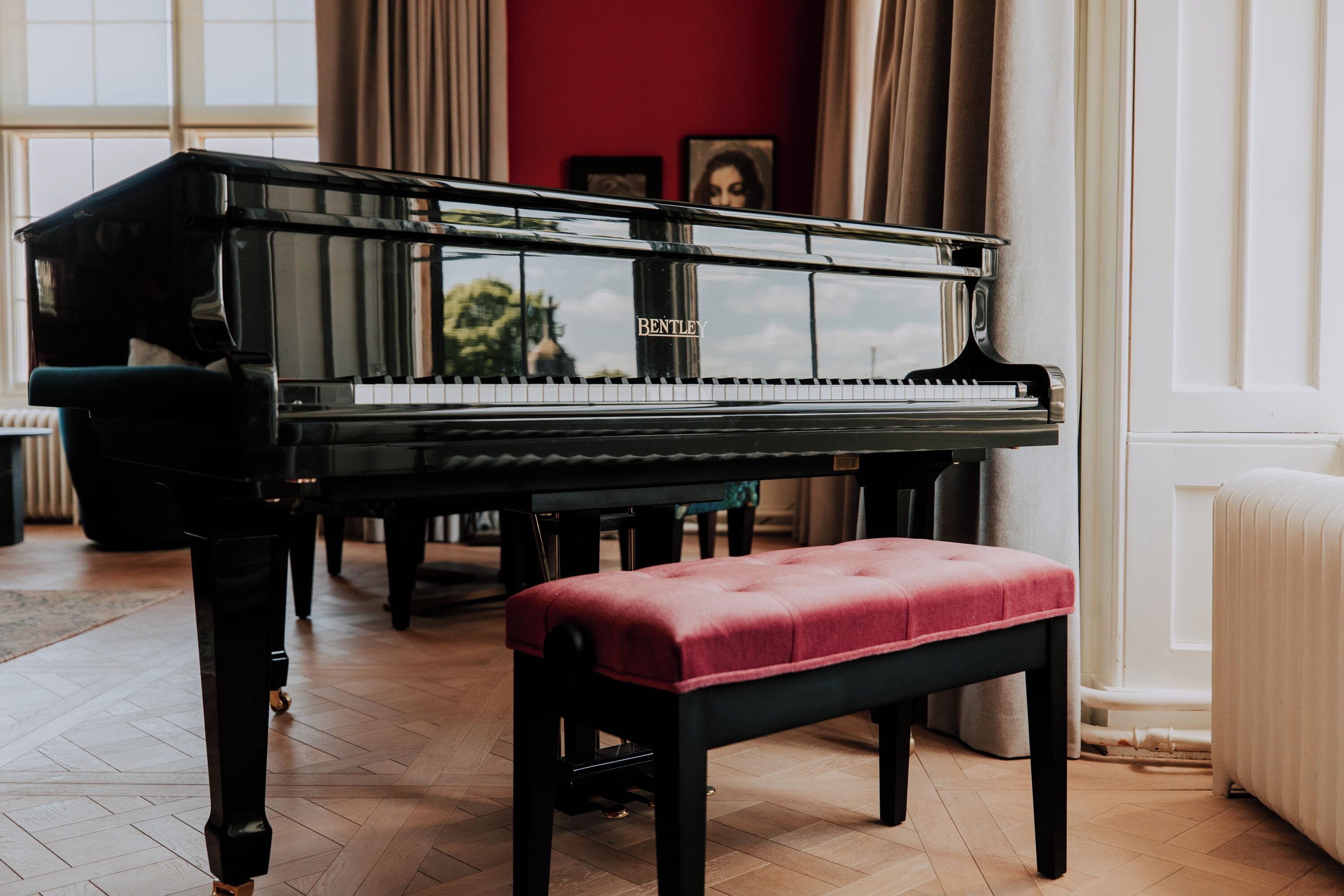 Een vleugelpiano in een kamer met een rode muur, een bankje en een raam.