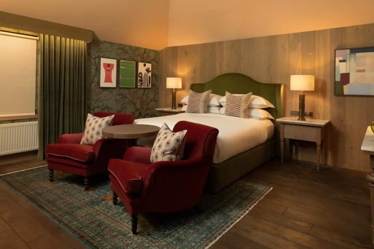 A hotel room with a bed, two red armchairs, a round table, a patterned rug, and two bedside lamps.