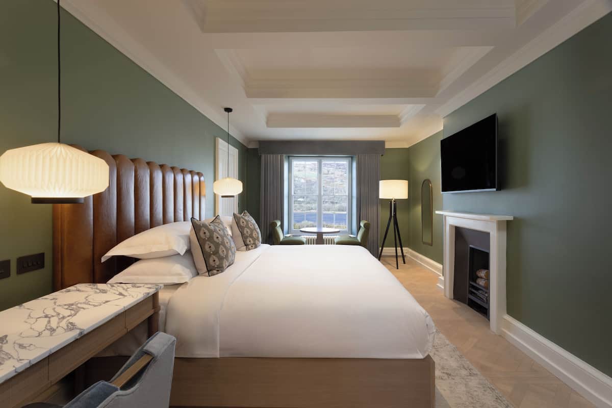A hotel room with a large bed, bedside tables, a desk, a TV, a fireplace, and a window with curtains.