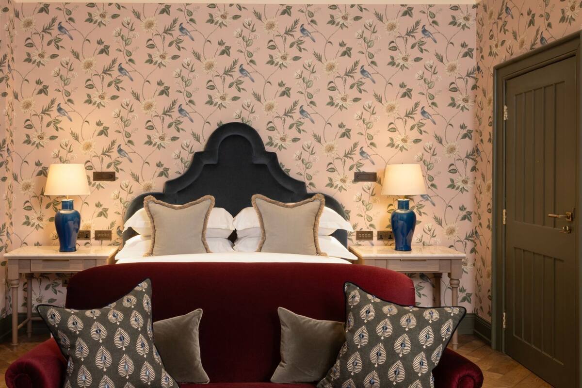 A bedroom with a large bed, two bedside tables with lamps, floral wallpaper, and a door.