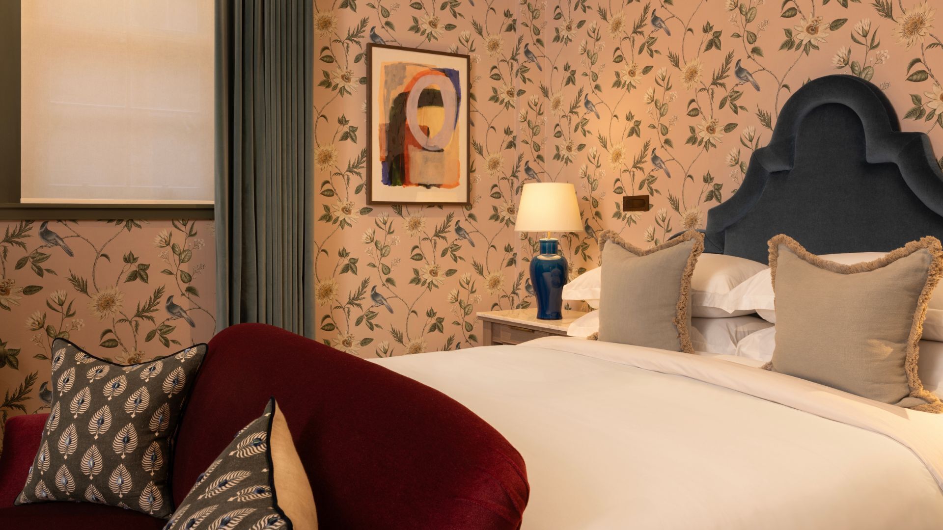 A hotel room with floral wallpaper, a bed with multiple pillows, a nightstand with a lamp, and a framed artwork on the wall.