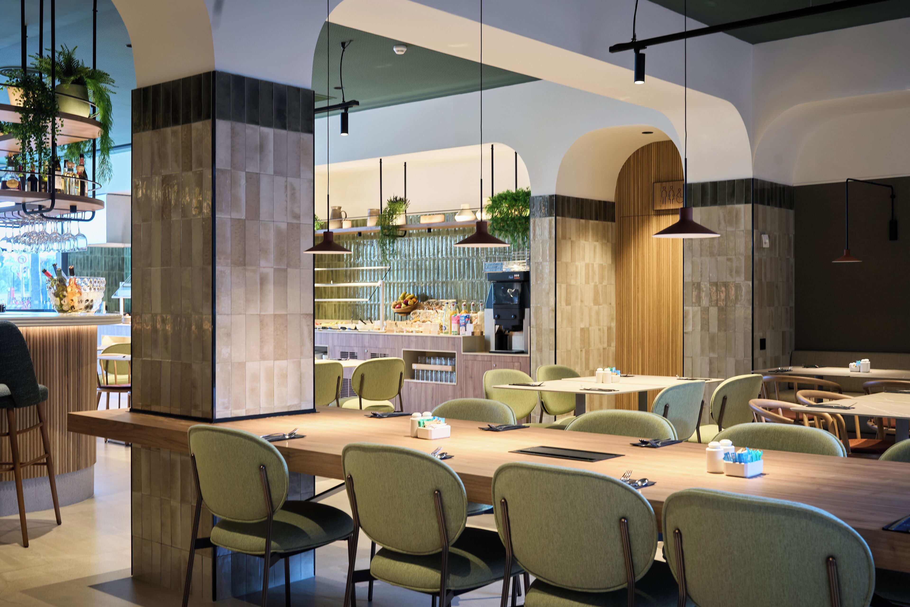 A modern restaurant with a long communal table, green chairs, and hanging pendant lights.