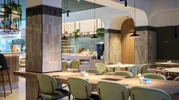 A modern restaurant with a long communal table, green chairs, and hanging pendant lights.