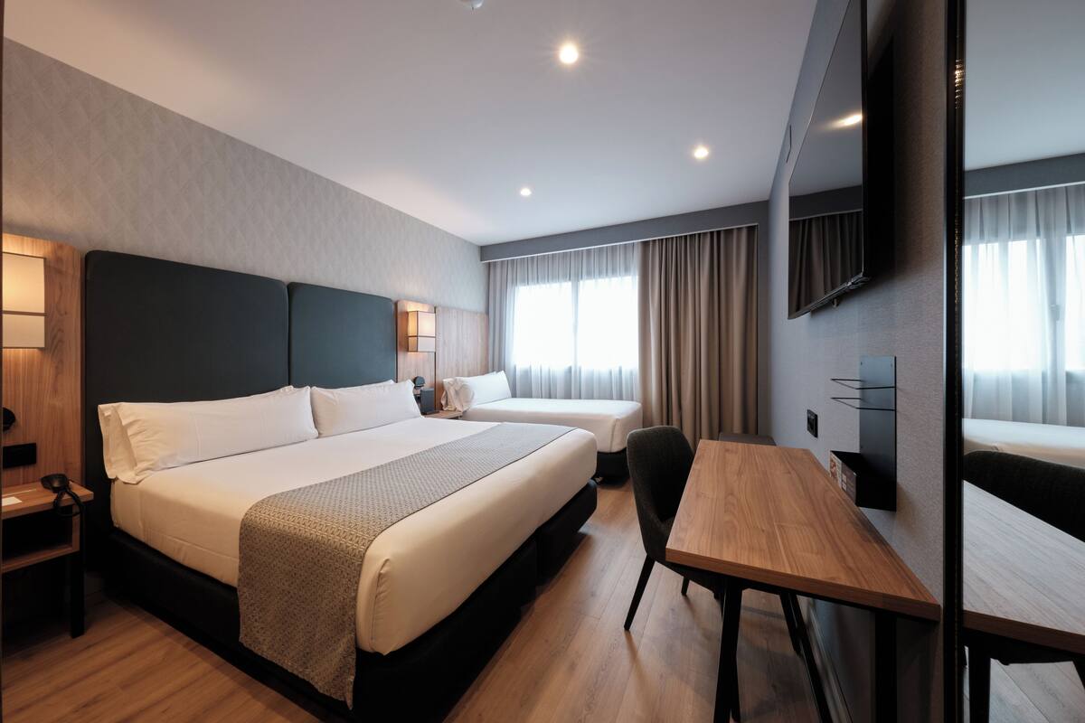 Une chambre d’hôtel avec un grand lit, un bureau, une chaise, une télévision et une fenêtre avec des rideaux.