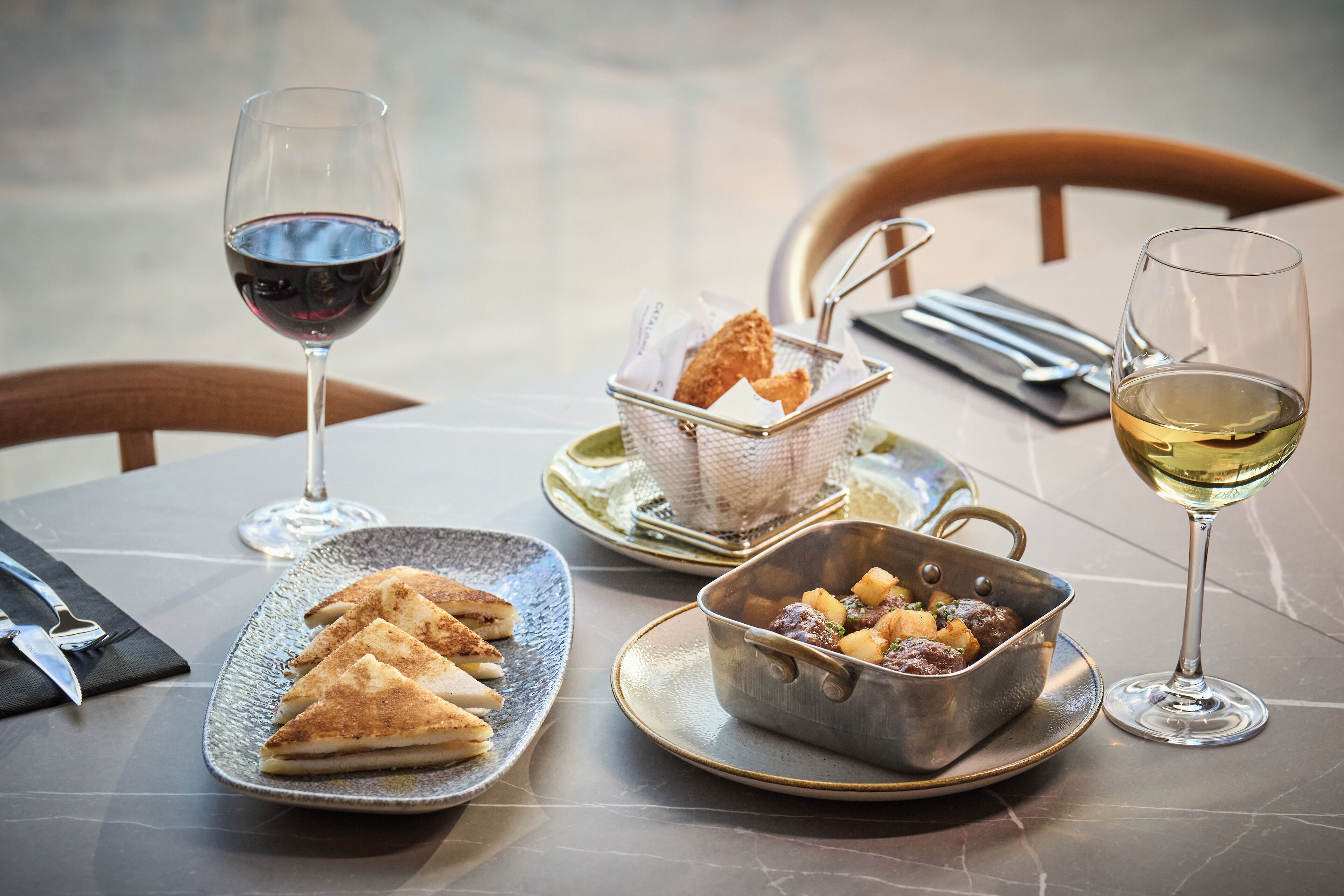A dining table with a plate of triangular pastries, a metal bowl with a stew, and two glasses of wine.