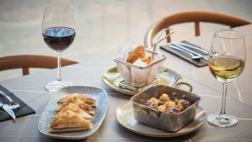 A dining table with a plate of triangular pastries, a metal bowl with a stew, and two glasses of wine.