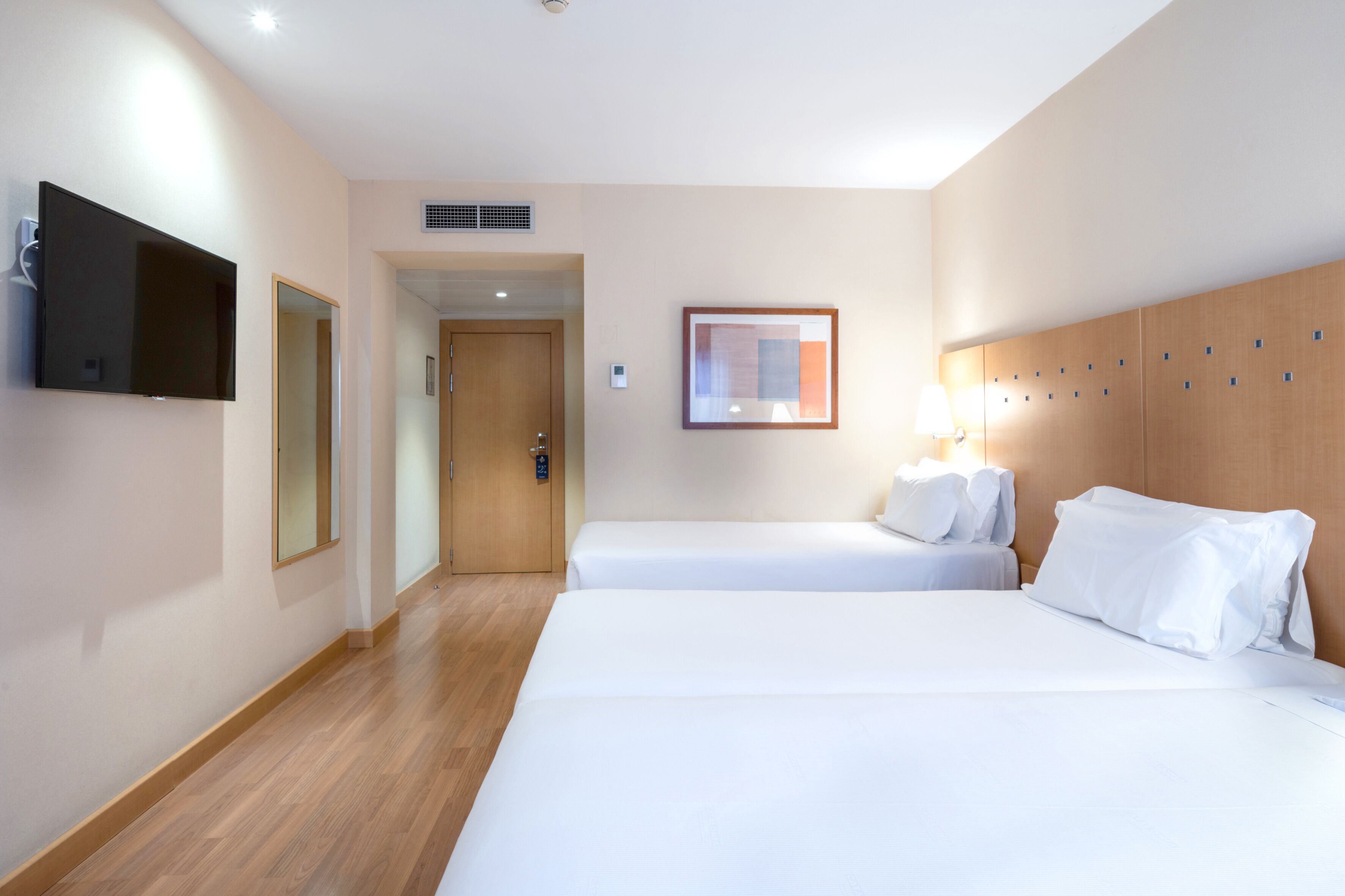 A hotel room with two single beds, a flat-screen TV mounted on the wall, a framed artwork above the bed, and a wooden headboard.