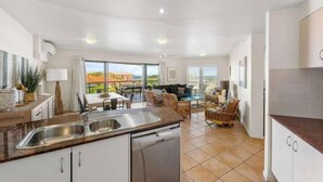 A modern kitchen with a dishwasher, a dining area with a table and chairs, and a view of the outdoors. - The Cove Yamba (Yamba)