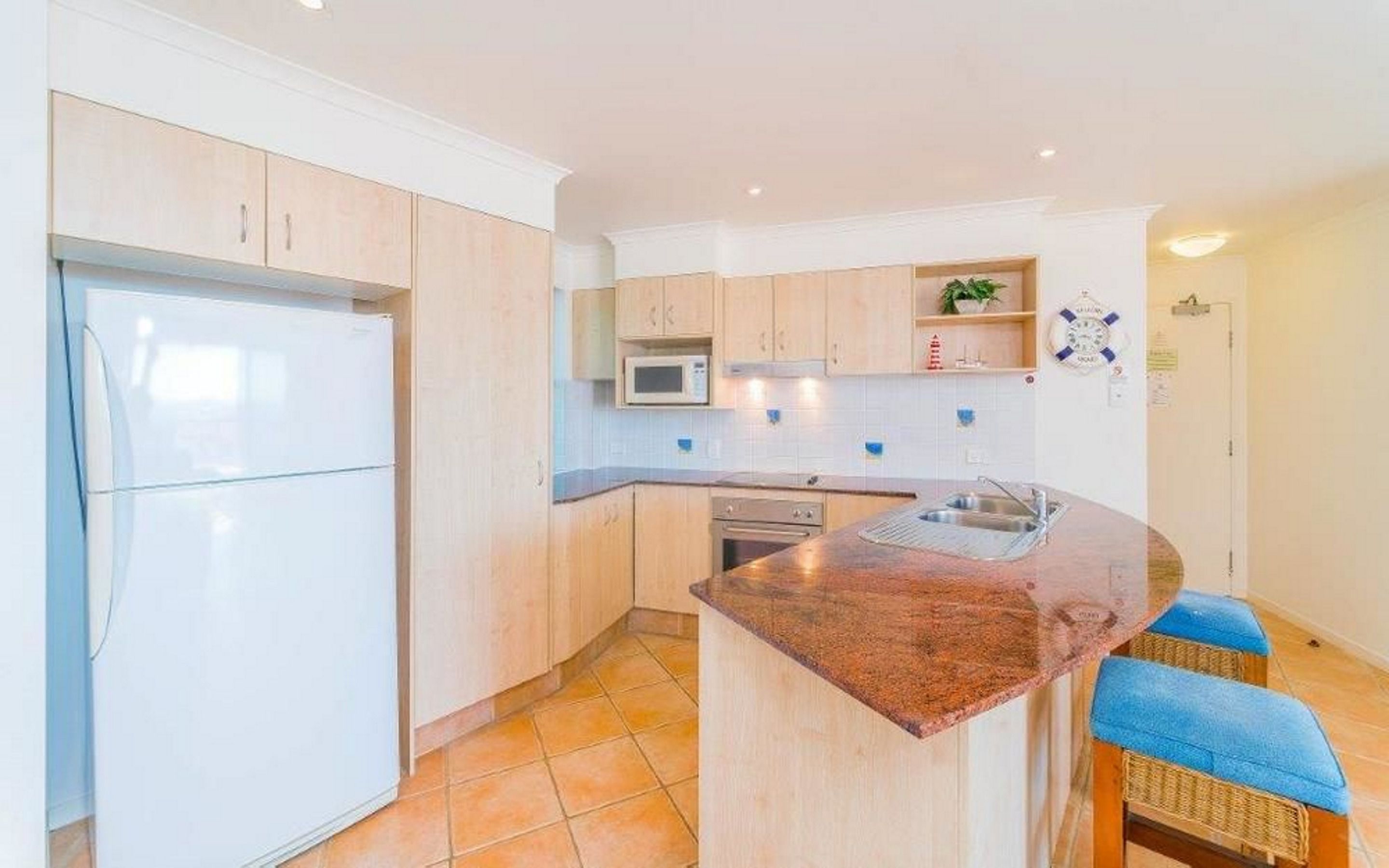 Een moderne keuken met lichte houten kastjes, een centraal kookeiland met barkrukken, een ingebouwde magnetron en een roestvrijstalen spoelbak.