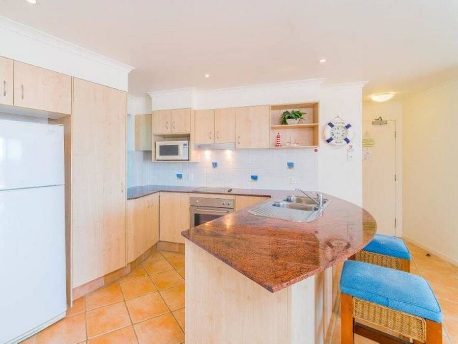 Een moderne keuken met lichte houten kastjes, een centraal kookeiland met barkrukken, een ingebouwde magnetron en een roestvrijstalen spoelbak.