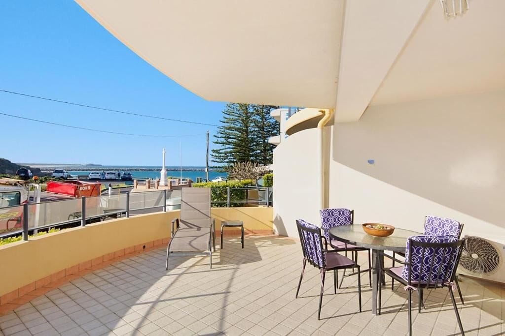 A balcony with a table and chairs, overlooking a coastal area with a view of the sea and a distant island.