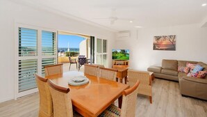 A spacious living room with a wooden dining table and wicker chairs, a sofa, and a television. - The Cove Yamba (Yamba)
