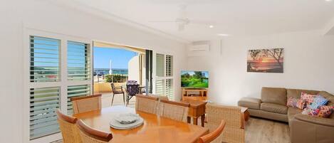 A spacious living room with a wooden dining table and wicker chairs, a sofa, and a television.