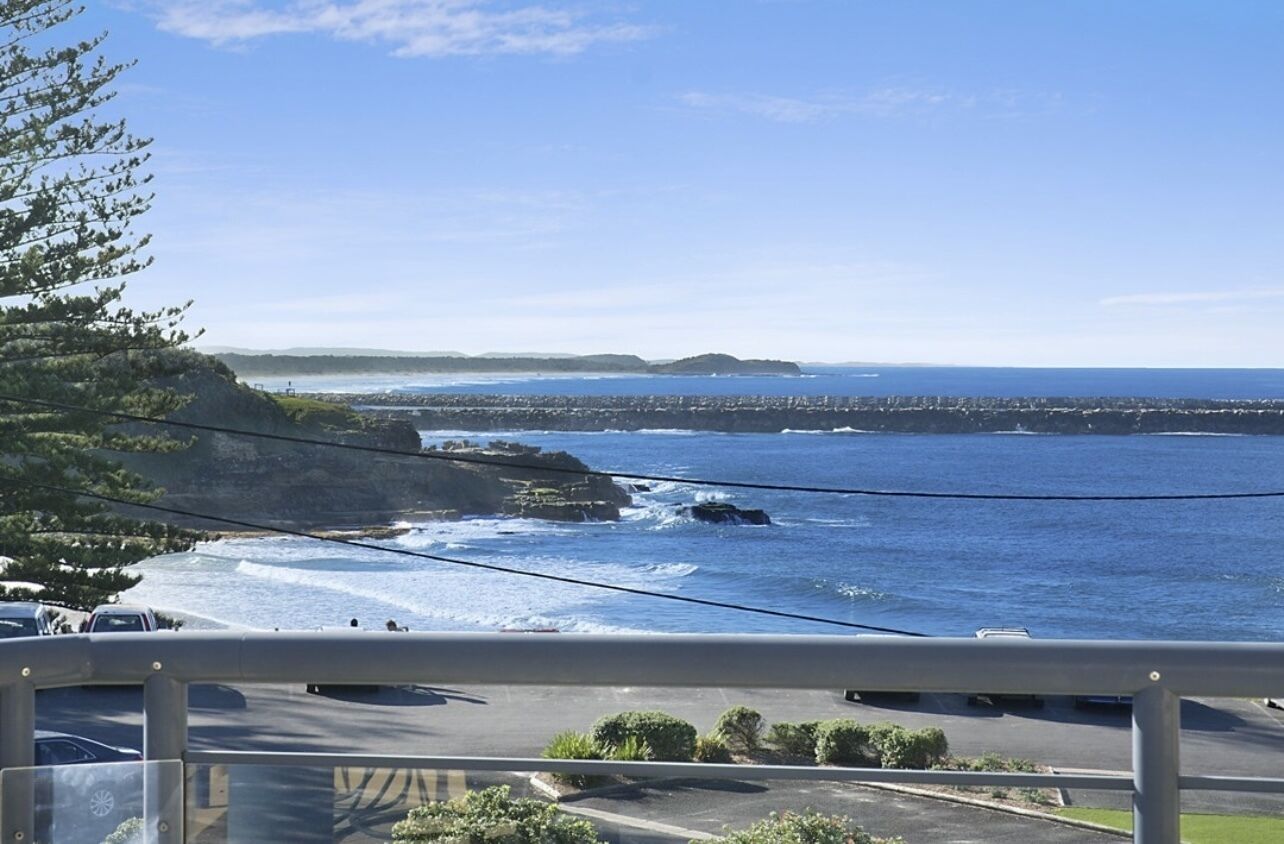A coastal view from a balcony with a clear sky, a rocky shoreline, and a distant cityscape.
