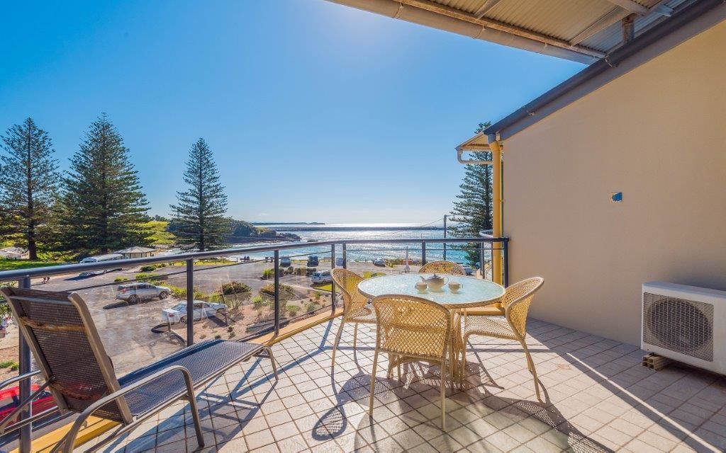 Un balcon avec une table et des chaises, donnant sur un parking et une étendue d’eau.