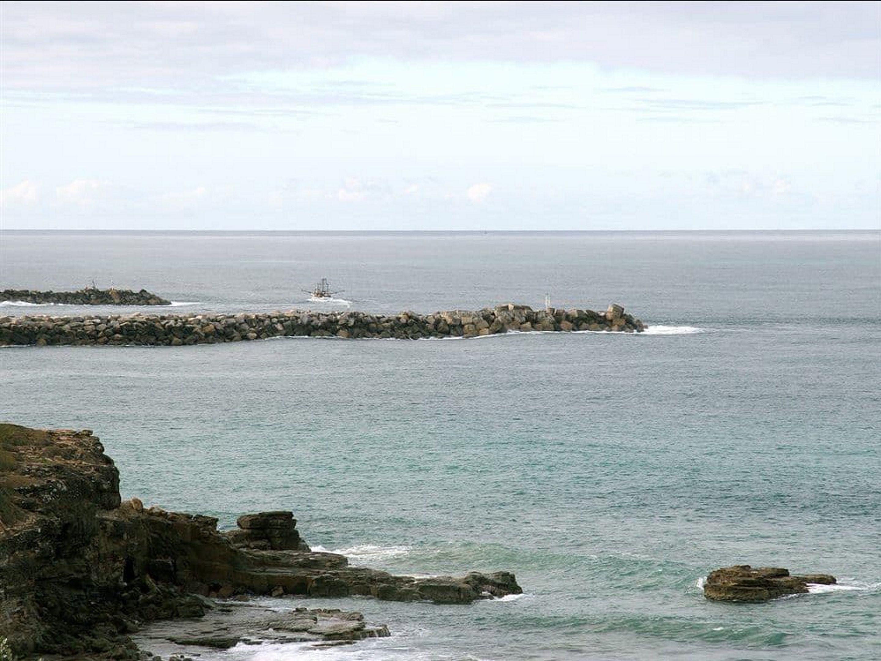 Un paisaje costero con un rompeolas rocoso que se extiende hacia el mar, un faro en el rompeolas y un litoral pedregoso en primer plano.
