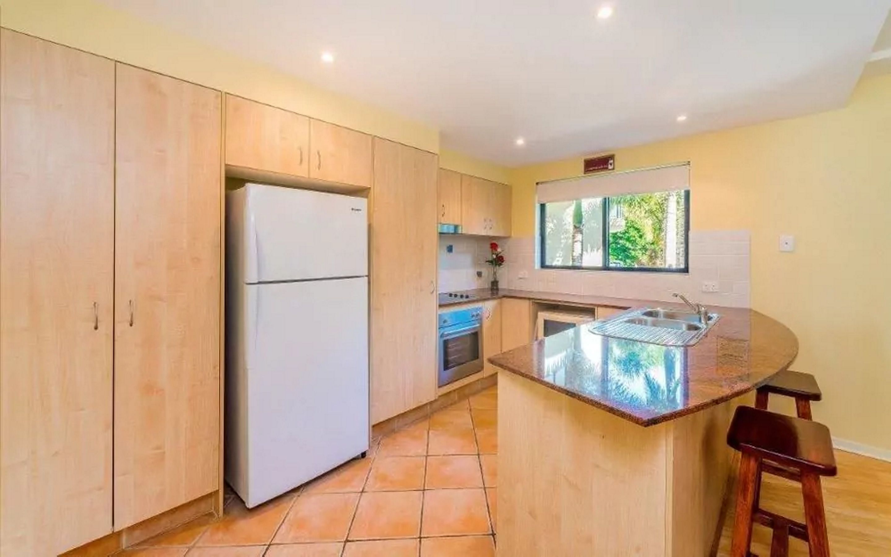 A compact kitchen with wooden cabinets, a built-in oven, a refrigerator, and a window providing natural light.