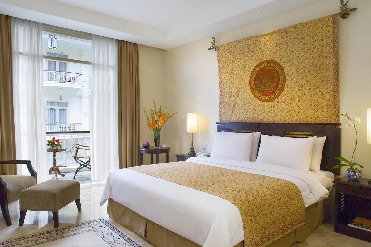 Ein Hotelzimmer mit einem großen Bett, einem kleinen Tisch mit Blumenvase, einem Sessel und Blick auf einen Balkon.