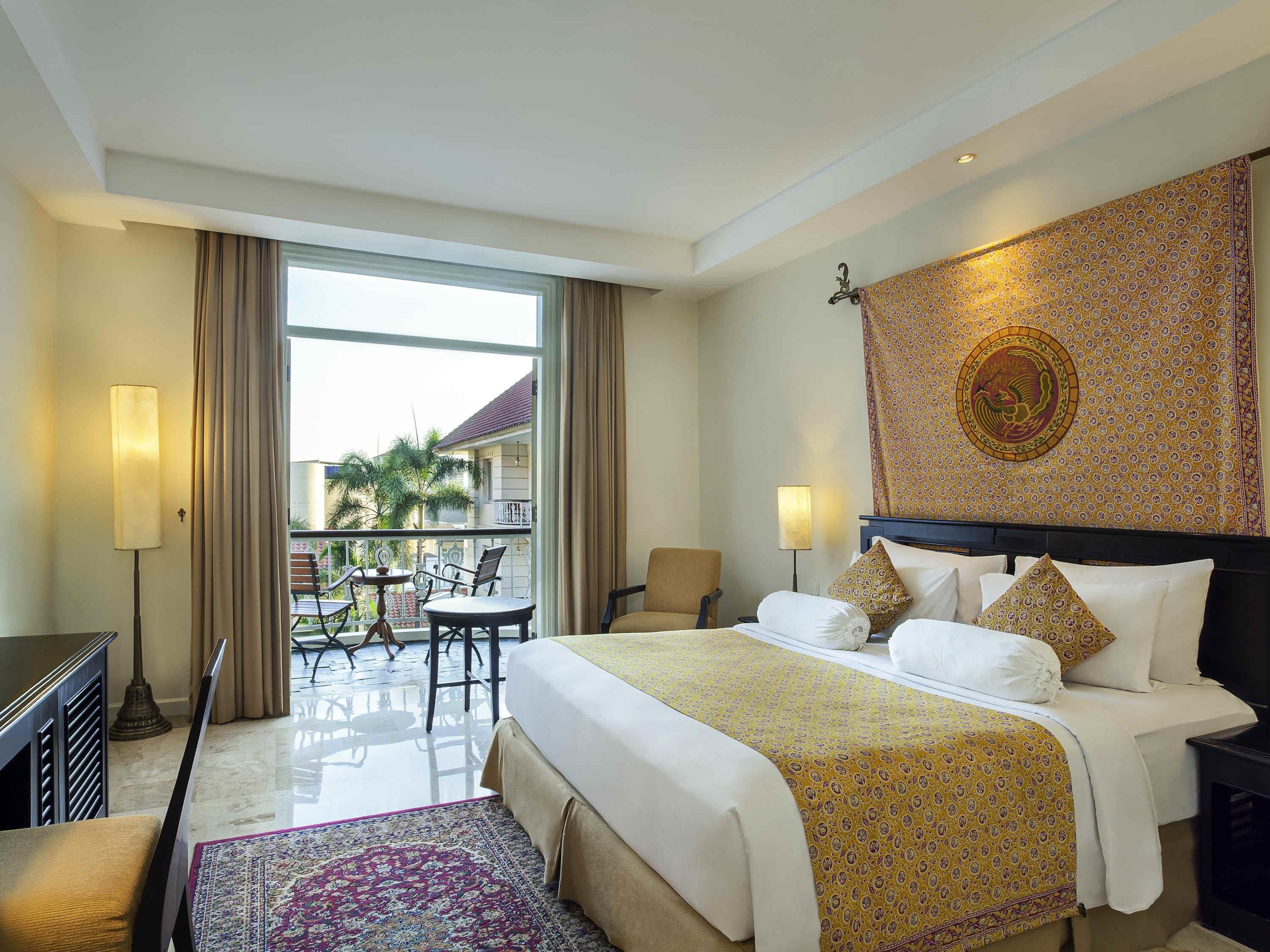 A hotel room with a bed, a desk, a chair, a balcony with a table and chairs, and a patterned wall tapestry.