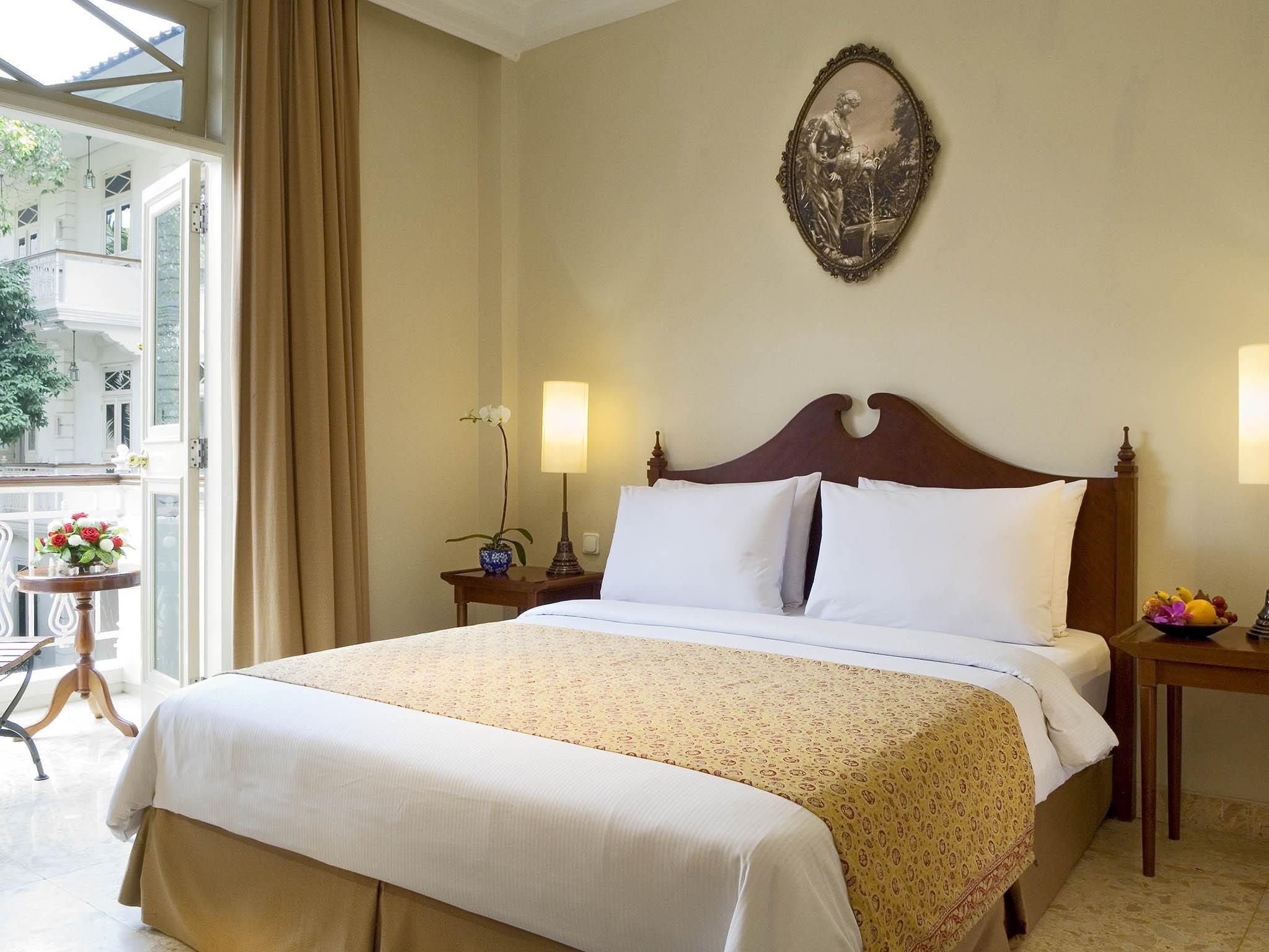 A hotel room with a large bed, two bedside tables, a nightstand, a window with curtains, a small table with a vase of flowers, and a wall-mounted picture.