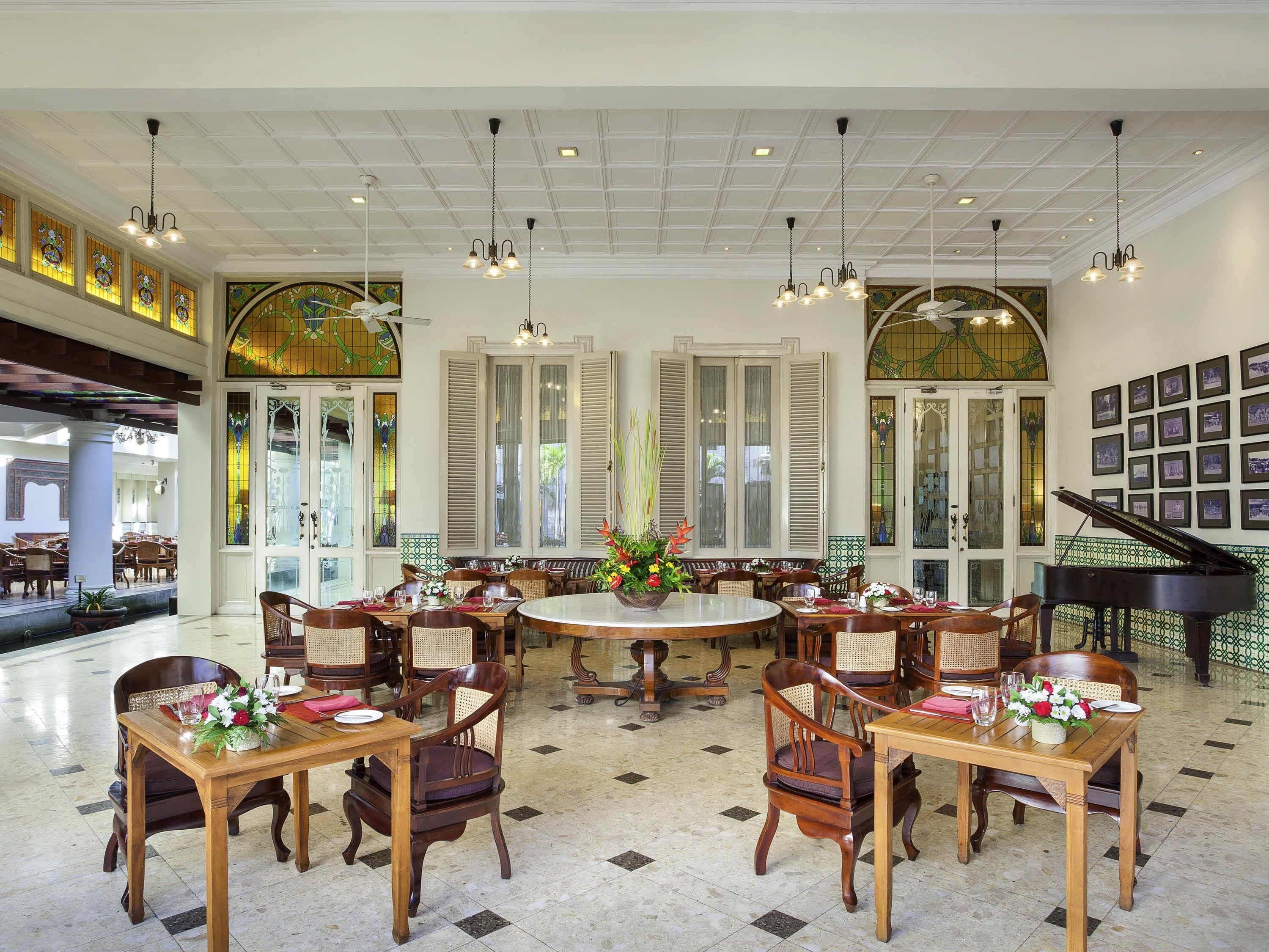 A spacious dining area with multiple tables and chairs, a grand piano, and large windows with shutters.