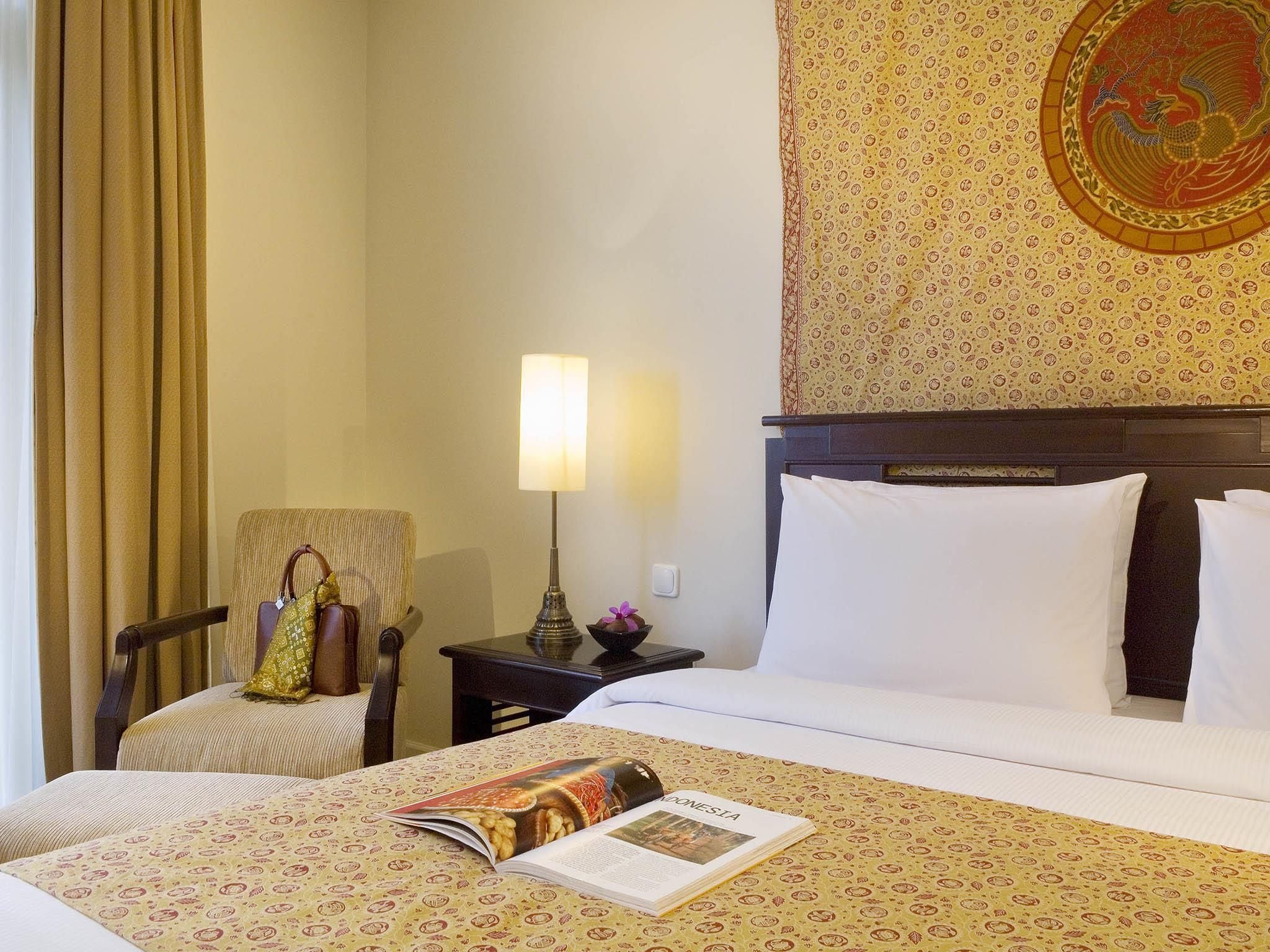 A hotel room with a bed, a chair, a nightstand, a lamp, and a wall with a decorative emblem.