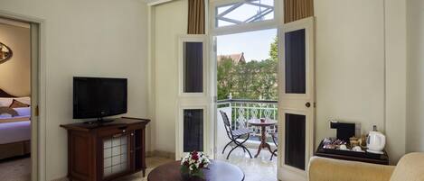 A hotel room with a TV, a sofa, a coffee table with flowers, a balcony with a table and chairs, and a view of greenery.