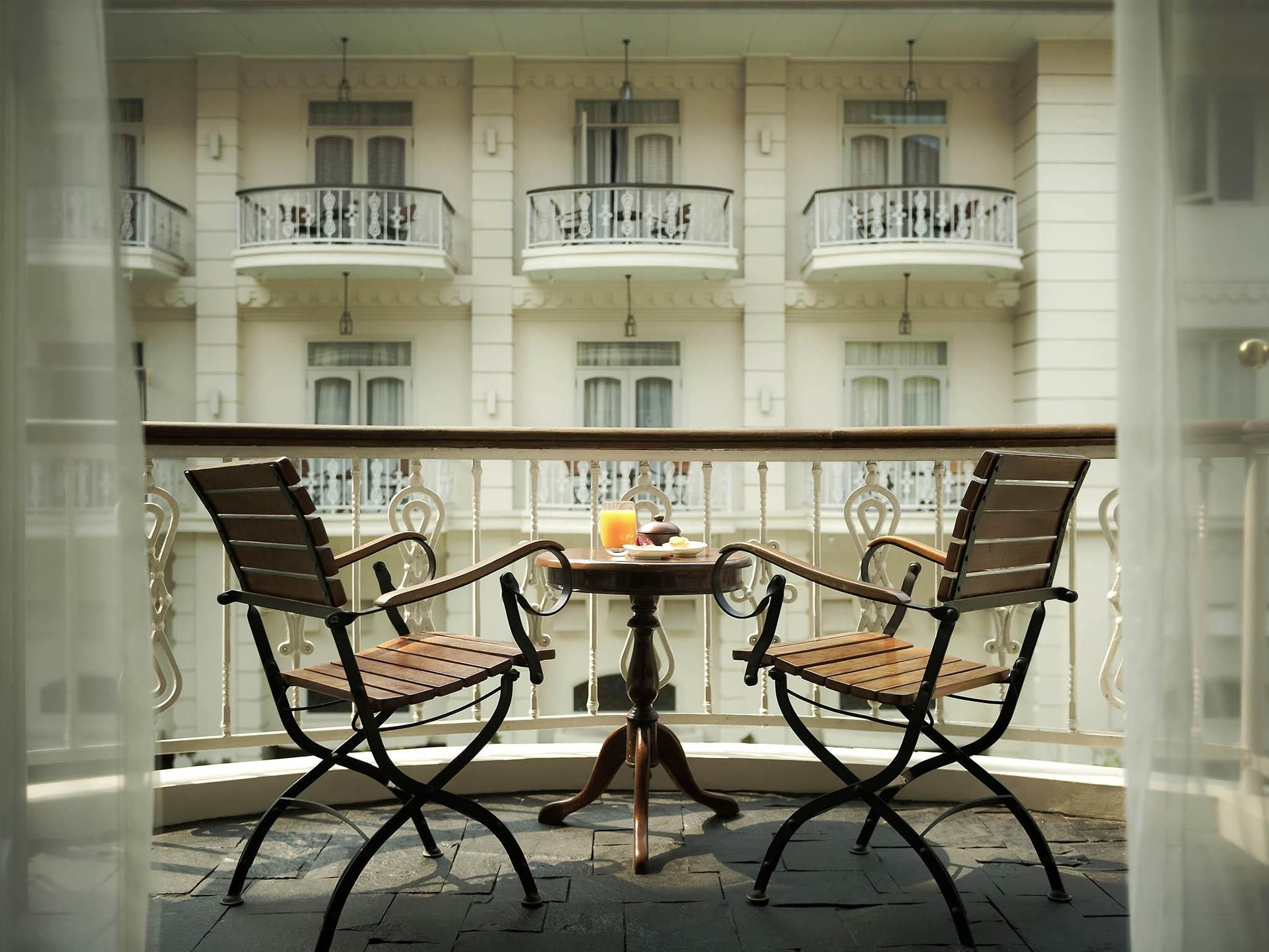 Een balkon met twee houten stoelen en een klein tafeltje, met uitzicht op een gebouw met meerdere balkons.
