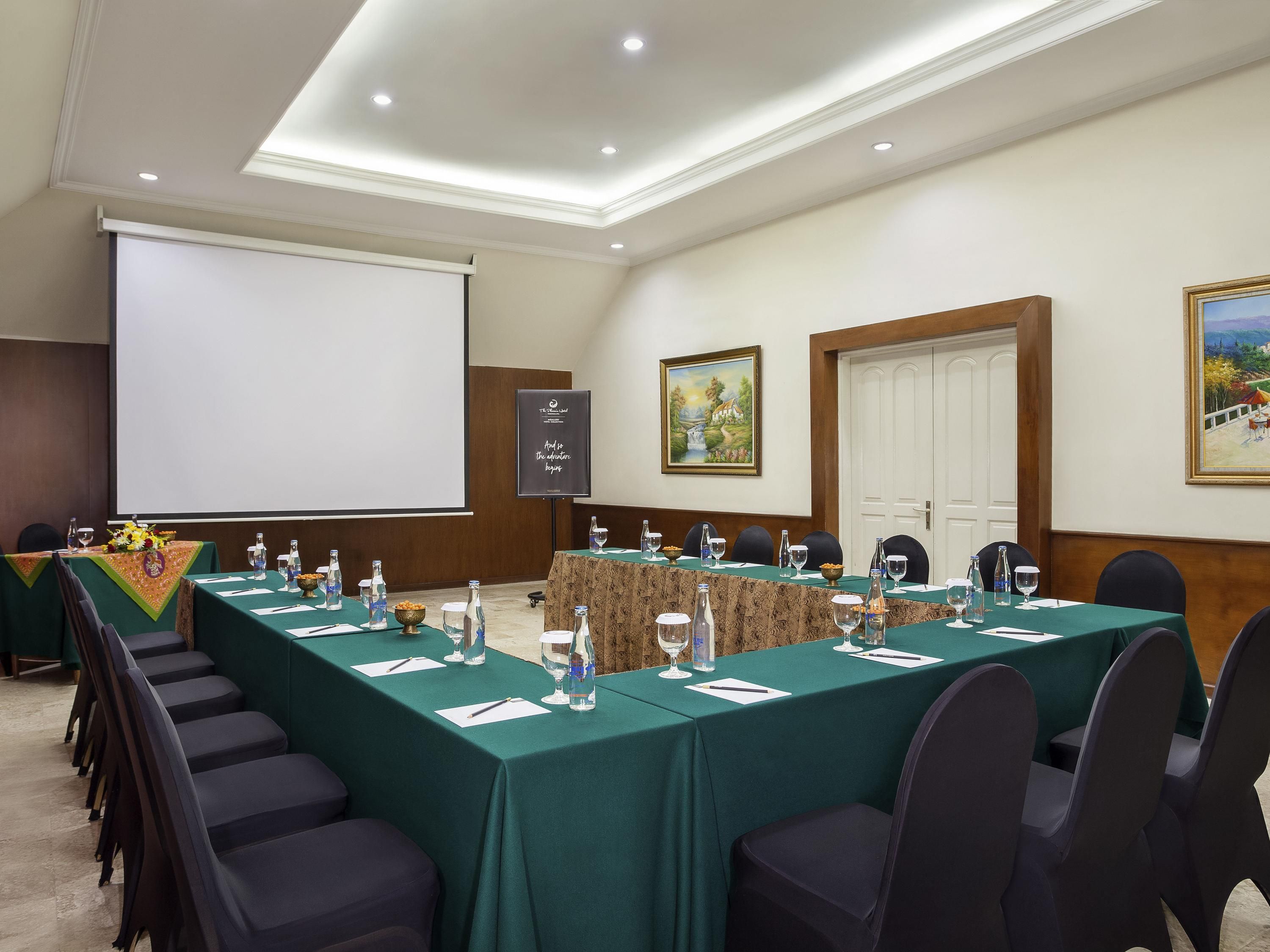 Un salón de conferencias con una larga mesa, sillas, una pantalla de proyector y un cuadro en la pared.