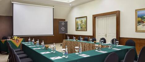 Un salón de conferencias con una larga mesa, sillas, una pantalla de proyector y un cuadro en la pared.