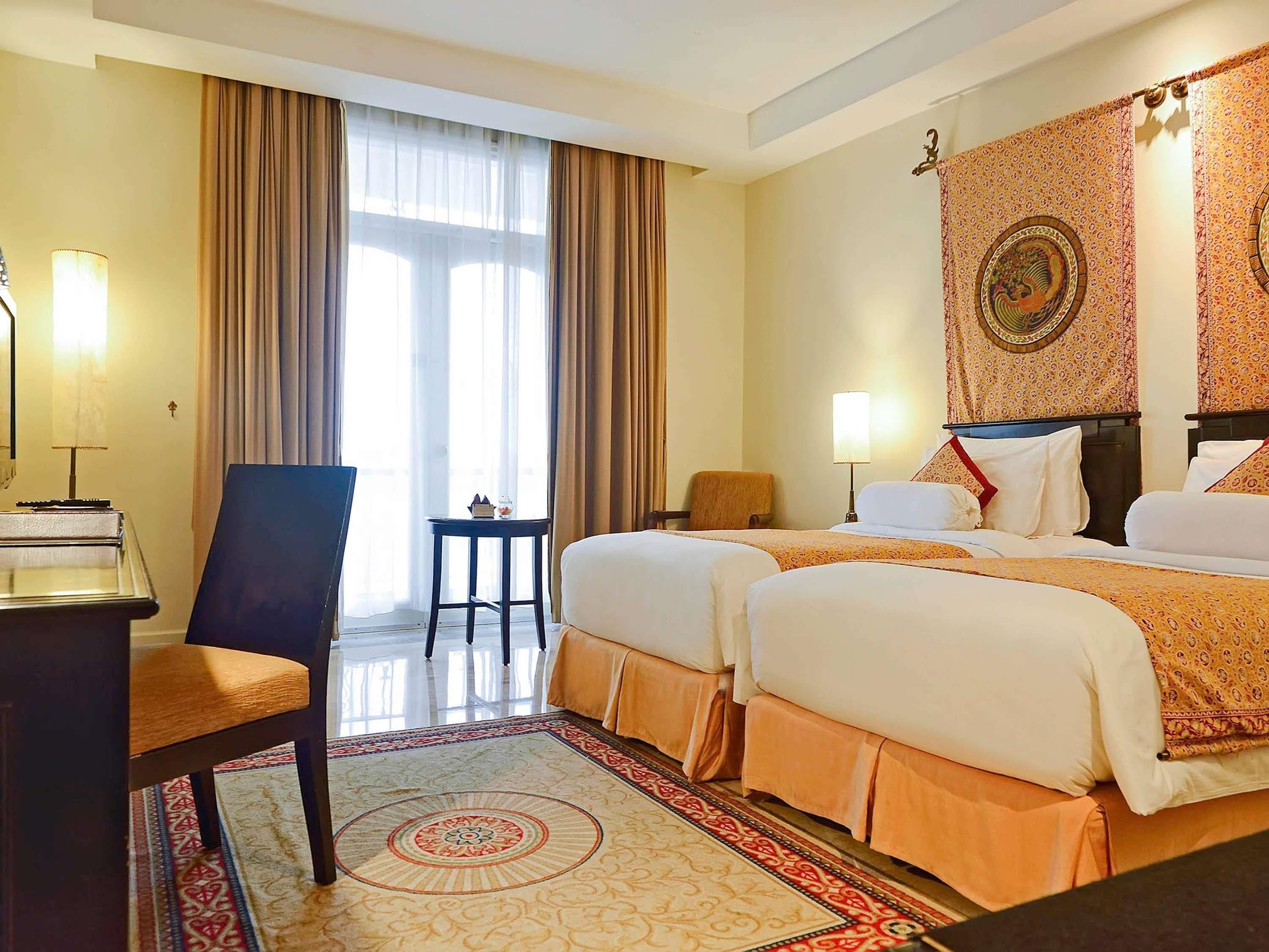 Ein Hotelzimmer mit zwei Betten, einem Schreibtisch, einem Stuhl, einem kleinen Tisch und einem großen Fenster mit Vorhängen.
