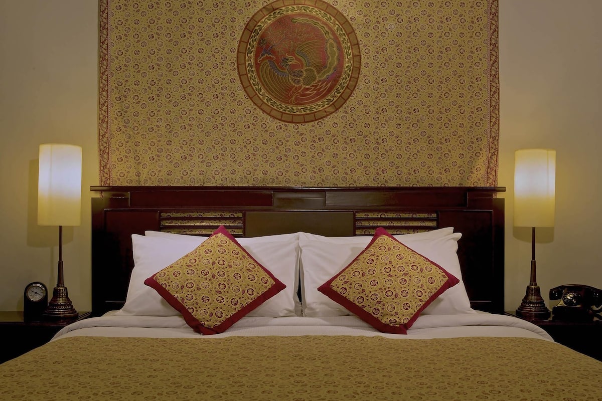 Habitación de hotel con un tapiz estampado, cabecera de madera, dos lámparas de noche y una cama con almohadas blancas y con diseño.