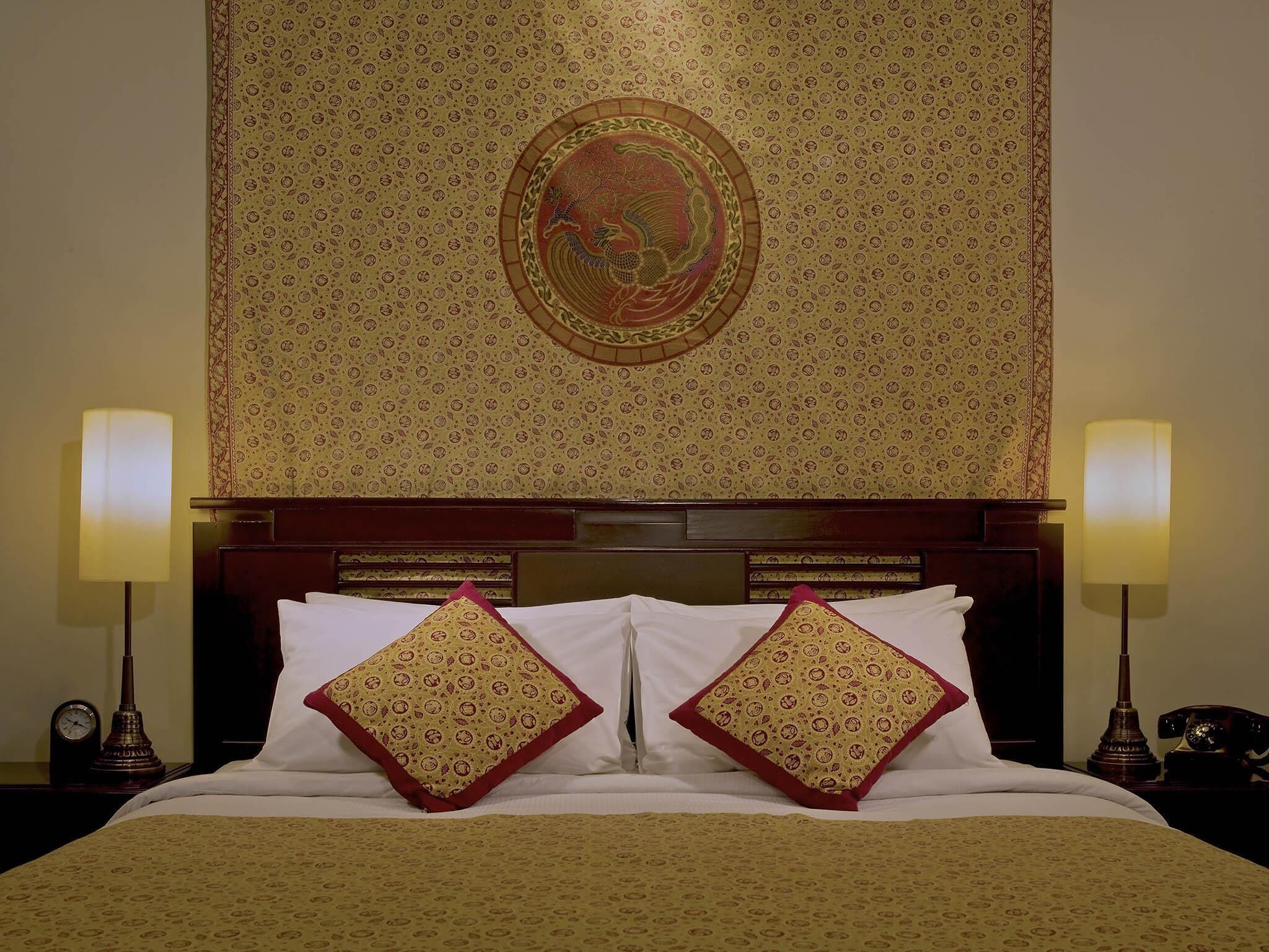 Een hotelkamer met een kamerkleed met patroon, een houten hoofdeinde, twee bedlampen en een bed met witte en gemêleerde kussens.