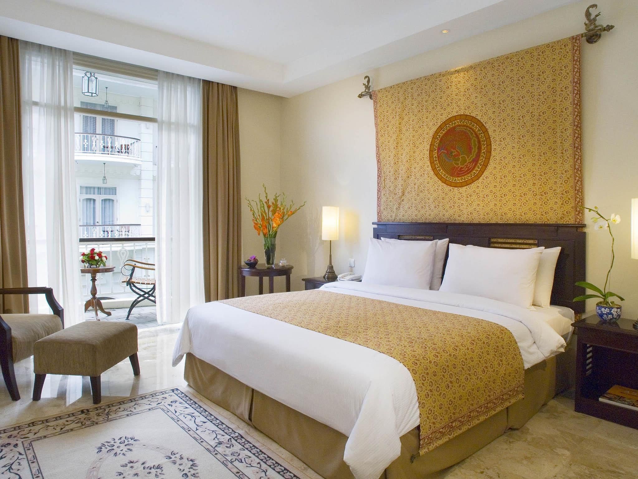 Habitación de hotel con una cama grande, una mesita con un florero, una silla y vistas a un balcón.
