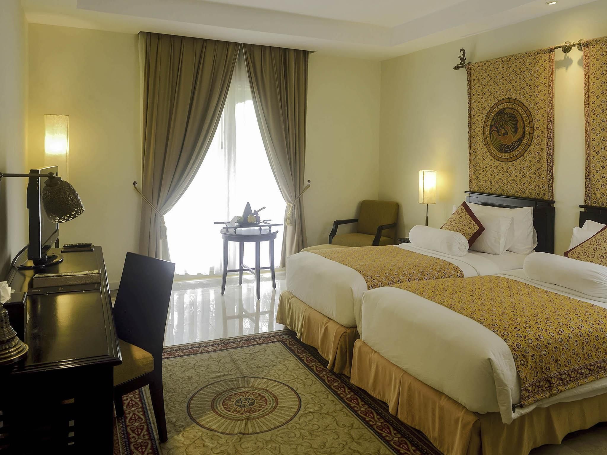 A hotel room with two beds, a desk with a lamp, a chair, a small table, and a large window with curtains.