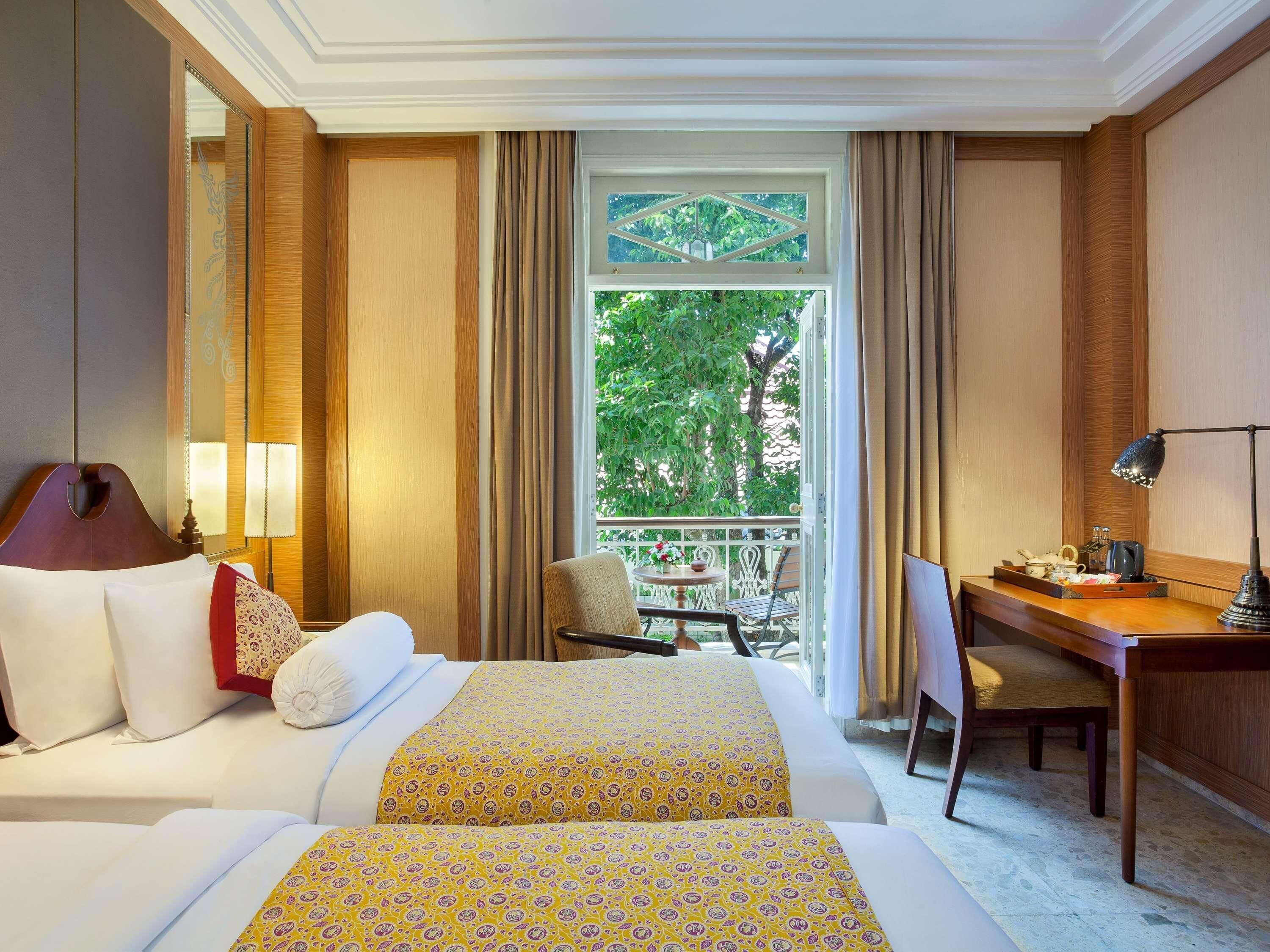 Habitación de hotel con dos camas, un escritorio, una silla, un balcón con vistas a árboles y una ventana con cortinas.