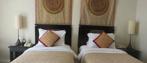 A hotel room with two single beds, each with a patterned bedspread and a decorative pillow. There are bedside tables with lamps and a telephone.