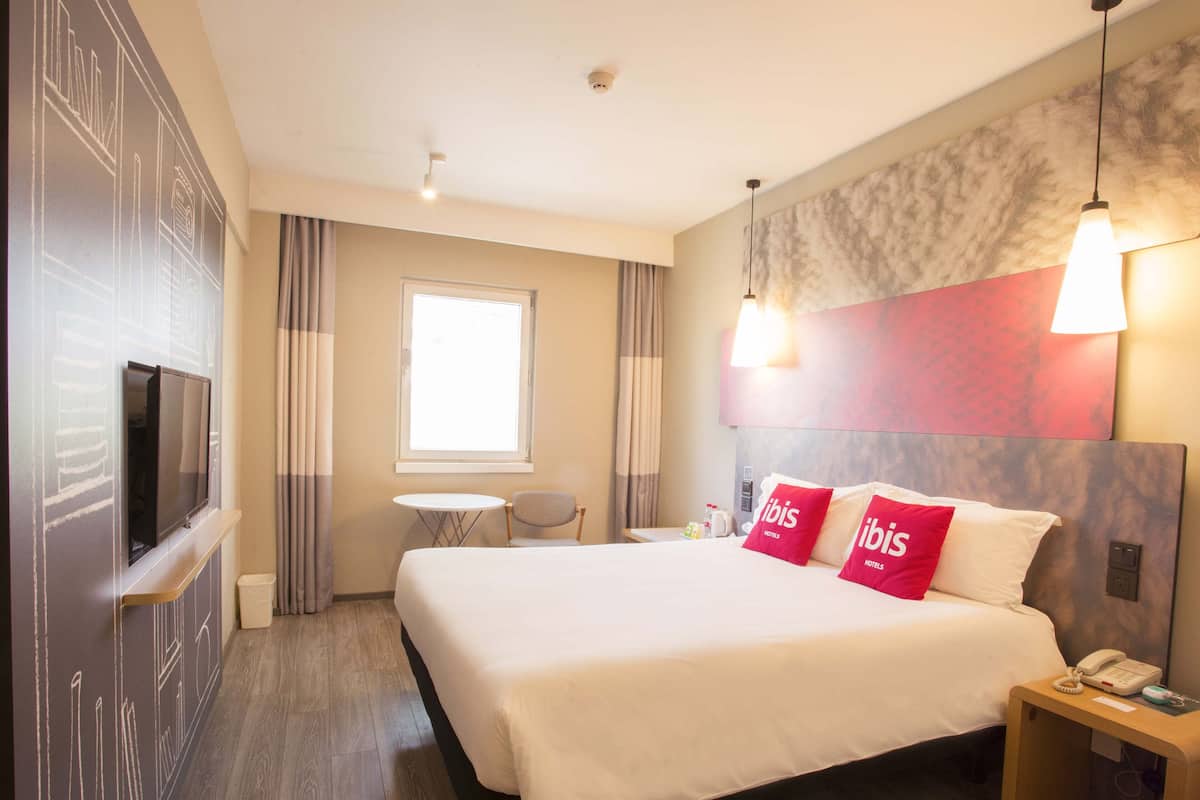 A hotel room with a large bed, a TV mounted on the wall, a small table, and a chair.