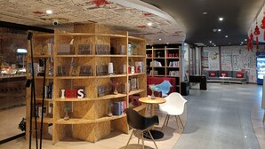 A modern interior with a wooden bookshelf displaying books and framed photos, a round table with chairs, and a seating area with red and blue chairs. - ibis Tianjing Teda (Tianjin)
